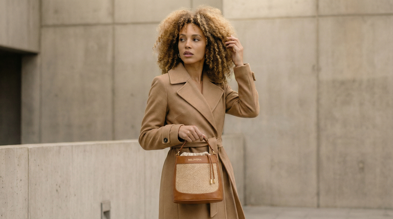 Woman wearing a beige coat in a concrete urban setting carrying the Milovena Vertical bag in cognac